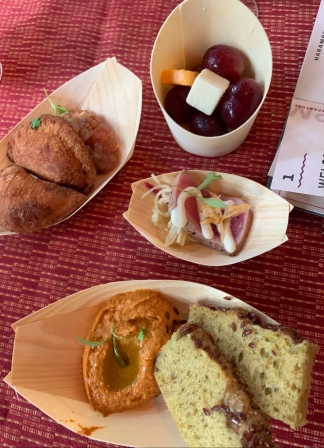 There is a red table cloth, atop are four plates. One has two cubes of cheese and four grapes, one with a slice of ahi tuna and white sauce, one with two pieces of bread an orange hummus, and one with two meat pies.