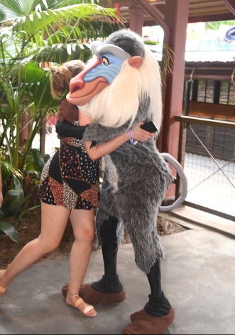 Writer is shown from the side in a black romper and sandals. Her face is hidden as she hugs Disney's Rafiki, a grey baboon.