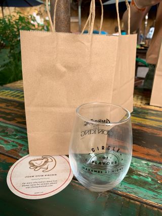 A brown paper bah sits on a wooden table with peeled green and yellow paint. In front of it is a transparent stemless wine glass that says "Circle of Flavors: Harambe at Night" on one side and "Celebration of the Lion King" on the other.