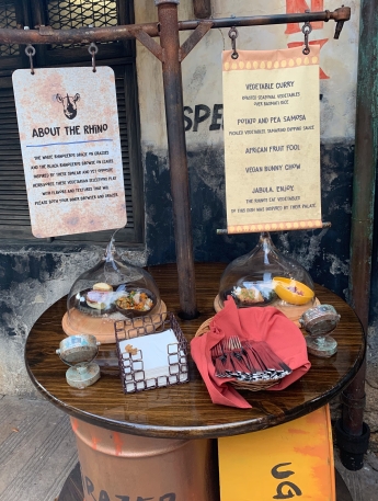 This is the display for the first station. There is a sign that lists each dish and a wooden table with napkins, silverware, and two display dishes under glass lids.