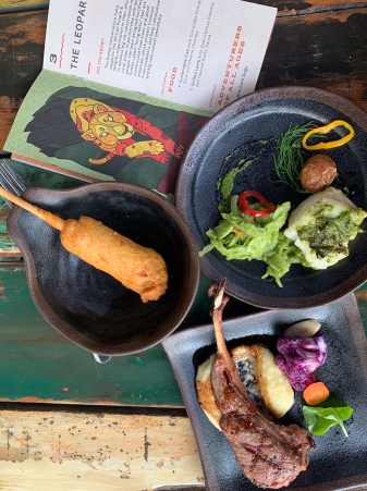 There are three plates. One has a corndog. The other has a large dollop of creamy polenta topped with a small leg of lamb with grill marks. There is also a piece of purple cauliflower on the side. The final plate has a piece of white fish covered in a green sauce. There is a small mini potato on the plate and a green slaw.