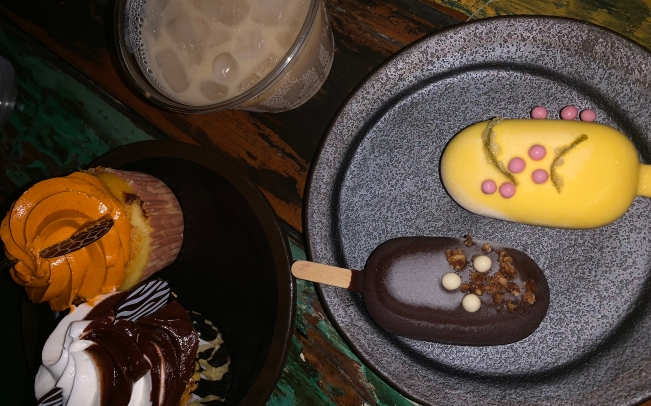 There are two plates. One has two cupcakes, one with orange frosting one with brown and white swirled frosting. The other plate has two frozen ice cream bars. One is dark chocolate dipped, the other in yellow-tinted white chocolate with pink candy pearls. And a beige alcoholic drink with ice.
