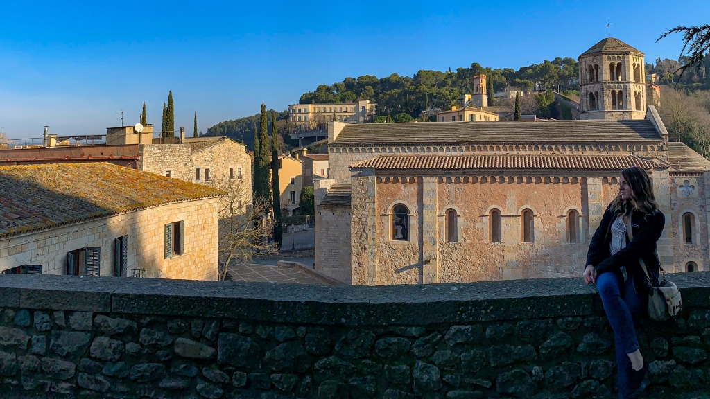 Writer sits on the right hand side of a short stone wall facing left with legs crossed. Behinds her are the roofs of a couple of Mediterranean buildings and a small tower to the right. Tall green trees stand behind the buildings.