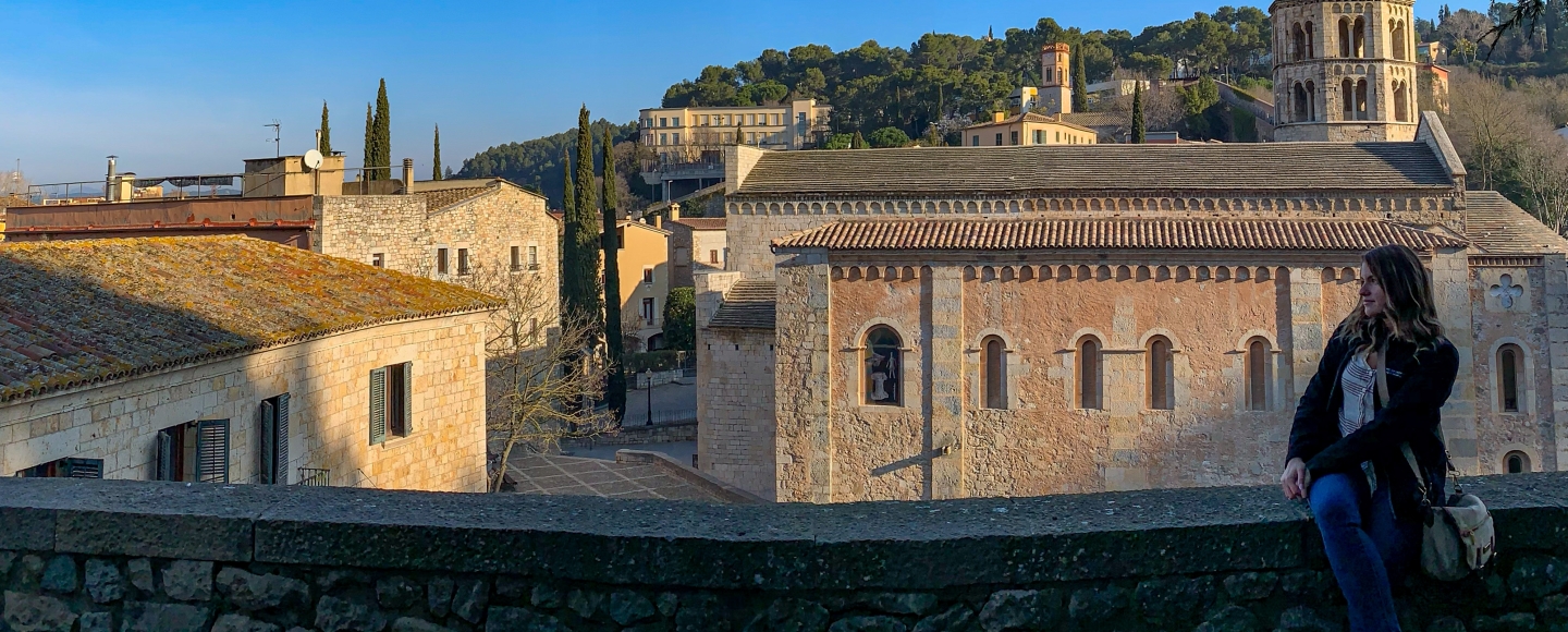 Writer sits on the right hand side of a short stone wall facing left with legs crossed. Behinds her are the roofs of a couple of Mediterranean buildings and a small tower to the right. Tall green trees stand behind the buildings.